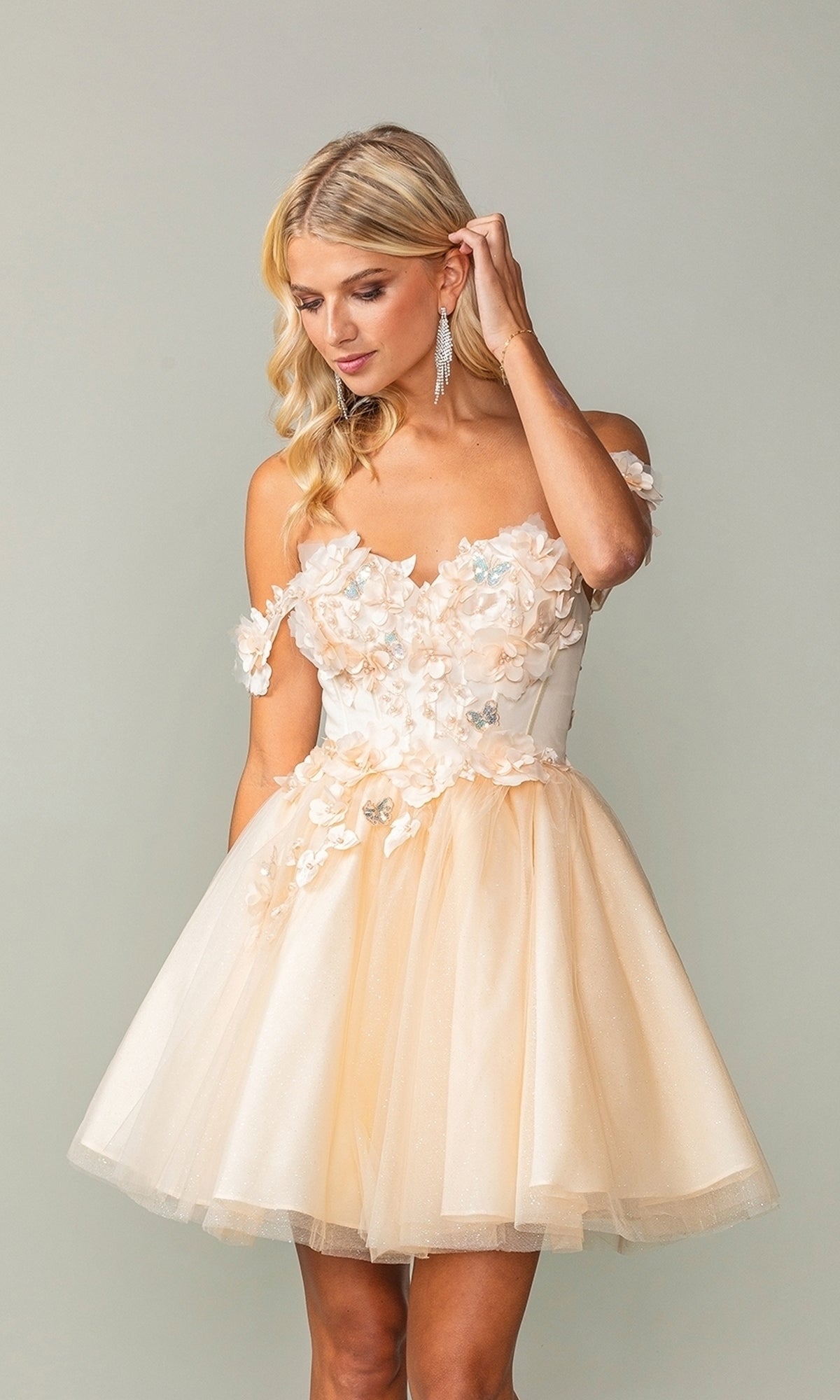 A woman in the Off-Shoulder Short Babydoll Homecoming Dress 3388, crafted from cream tulle with floral appliqués, stands against a plain backdrop and gently touches her hair.